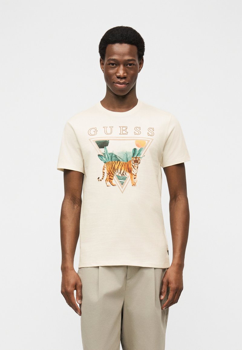 Άνδρας που φοράει μπεζ μπλουζάκι με γραφικό τίγρης και τροπικών φύλλων κάτω από τη λέξη "GUESS" και παντελόνι ανοιχτού χρώματος.