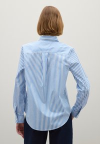 Personne avec des cheveux châtains clairs courts portant une chemise à rayures verticales bleues et blanches à manches longues, vue de dos sur un fond uni.