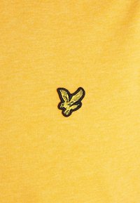 Camiseta de algodón amarilla con un logo de ave texturizado y bordado en negro y amarillo en el pecho. Tejido suave y diseño casual.