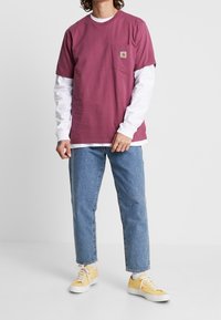 Weinrotes kurzärmliges T-Shirt mit einer Brusttasche, darüber ein weißes langärmliges Shirt, kombiniert mit hellblauem Jeans und gelben Turnschuhen.