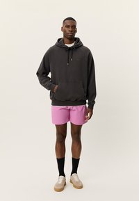 Μαύρο φούτερ με κουκούλα, τσέπη καγκουρό και ρυθμιζόμενα κορδόνια, συνδυασμένο με ροζ υφασμένα σορτς. Υποδήματα: μπεζ αθλητικά παπούτσια με άσπρες σόλες.