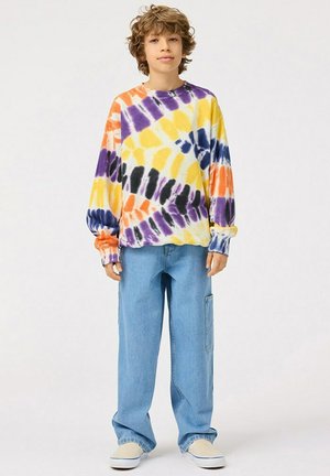 Maglietta a manica lunga tie-dye con motivi viola, giallo, arancione e nero. Abbinata a pantaloni di jeans blu larghi e scarpe di colore chiaro.