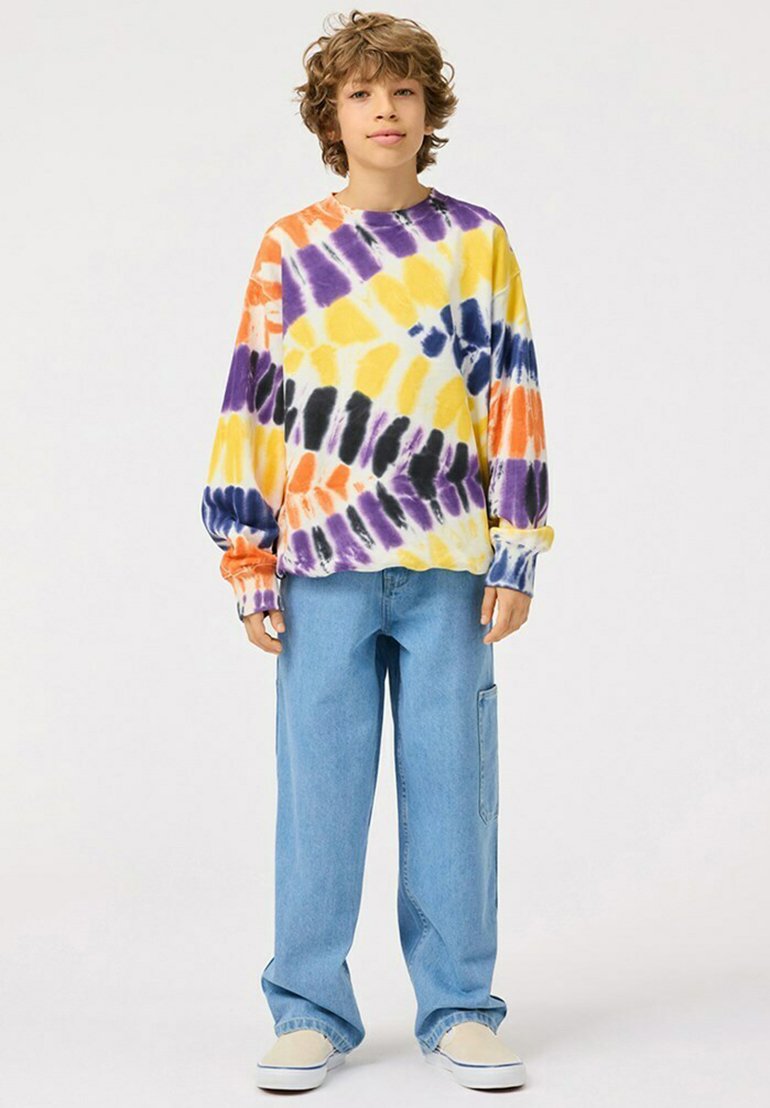 Maglietta a manica lunga tie-dye con motivi viola, giallo, arancione e nero. Abbinata a pantaloni di jeans blu larghi e scarpe di colore chiaro.