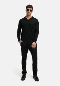 Schwarzer Strickpullover mit V-Ausschnitt und langen Ärmeln, kombiniert mit schwarzen Slim-Fit-Hosen und schwarzen Sneakers; das Modell trägt dunkle Sonnenbrillen.