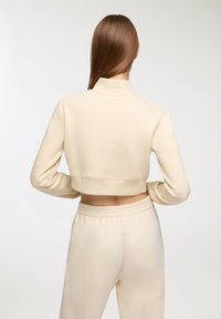 Felpa corta di colore beige chiaro con colletto alto e maniche lunghe, realizzata in tessuto morbido. Abbinata a pantaloni beige coordinati.