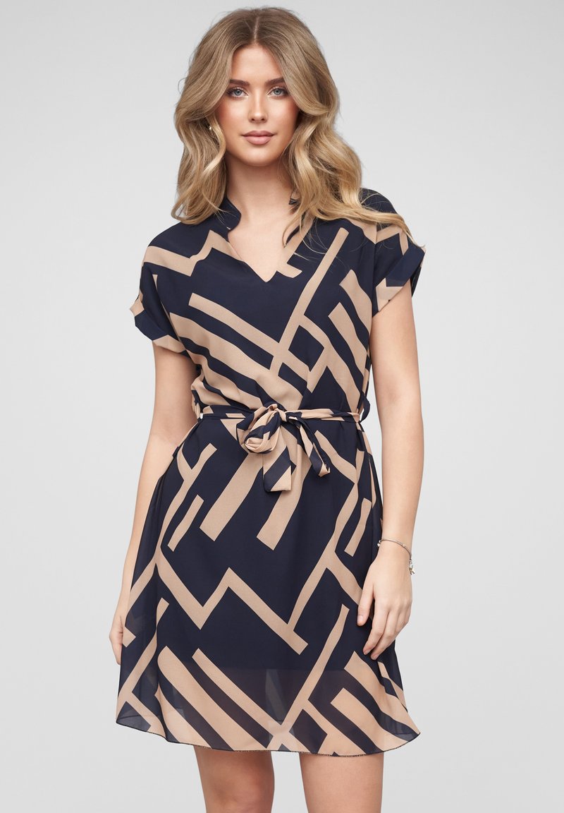 Femme aux cheveux blonds portant une robe bleu marine à manches courtes avec des lignes géométriques beiges et une ceinture nouée à la taille, debout devant un fond clair uni.