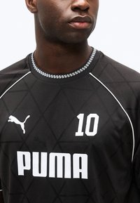 Άνδρας που φοράει μαύρη αθλητική φανέλα Puma με αριθμό 10, διακοσμημένη με λευκή διακόσμηση και λαιμόκοψη στρογγυλή με σχέδιο.