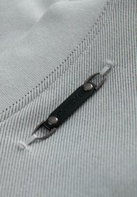 Primo piano di un tessuto grigio chiaro con un cinturino in gomma nera impresso con la parola "ambre", fissato da due clip metalliche che si attaccano a anelli in tessuto.