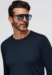 Blaue Sonnenbrille mit quadratischem Rahmen und getönten Gläsern, kombiniert mit einem marineblauen Strickpullover mit geripptem Rundhalsausschnitt und langen Ärmeln.