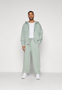 Ljusgrön hoodie och matchande träningsbyxor i mjukt tyg. Hoodien har dragkedja framtill och två fickor, med en avslappnad passform. Vit t-shirt under.