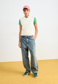 Gilet a trecce di colore crema su una T-shirt verde, jeans blu rilassati e sneakers verdi con dettagli bianchi, posati su un pavimento giallo.