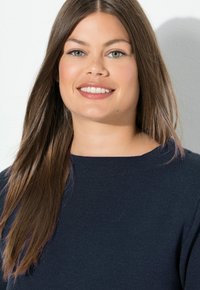 Model trägt einen dunkelblauen Strickpullover mit Rundhalsausschnitt, hat lange, gerade braune Haare und einen hellen Hintergrund.