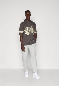Tricou sportiv gri cu un logo inspirat din cultura nativilor americani, accente crem și mâneci scurte, asociat cu pantaloni cargo gri deschis și adidași albi.