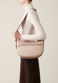 Borsa a spalla texturizzata rosa chiaro con forma curvata, dotata di una zip dorata e una tracolla marrone. Indossata a tracolla, abbinata a un top chiaro.