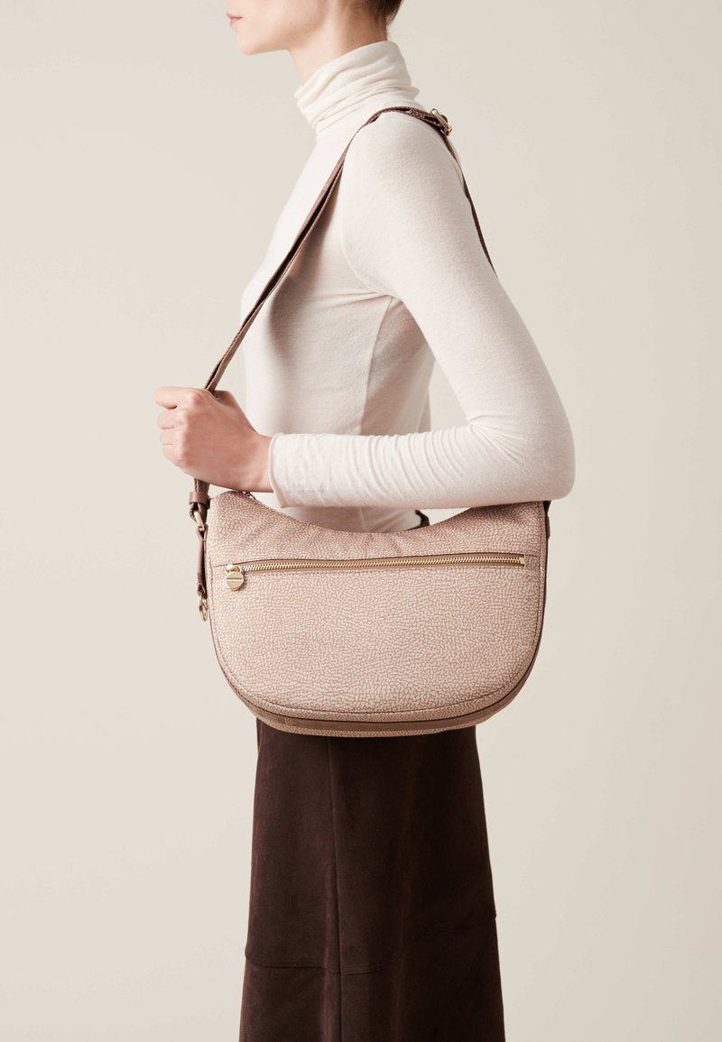 Borsa a spalla texturizzata rosa chiaro con forma curvata, dotata di una zip dorata e una tracolla marrone. Indossata a tracolla, abbinata a un top chiaro.