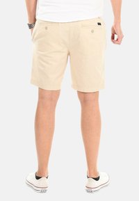 Beige Baumwollshorts mit geradem Schnitt, aufgesetzten Taschen mit Knöpfen und umgekrempeltem Saum, kombiniert mit weißen Sneakern vor einem hellen Hintergrund.