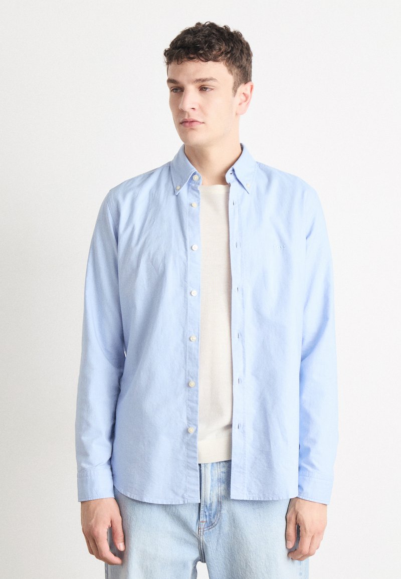 Lichtblauw langemouw shirt van katoen, met een button-down kraag en witte knopen, gelegd over een crèmekleurig top.