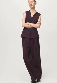 Fioletowy top bez rękawów z dekoltem w serek i falbaną, zestawiony z szerokimi spodniami wykonanymi z fakturowanego materiału, noszony z czarnymi butami.
