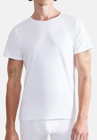 Weißes Baumwoll-T-Shirt mit kurzen Ärmeln, Rundhalsausschnitt, weicher Textur und tailliertem Schnitt. Zeigt sichtbare Tattoos an beiden Armen.