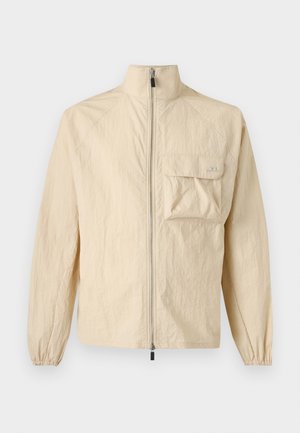 Homme portant une veste à fermeture éclair beige avec poche poitrine, short assorti et casquette, chaussettes noires, baskets, tenant un sac fourre-tout noir dans la main droite.