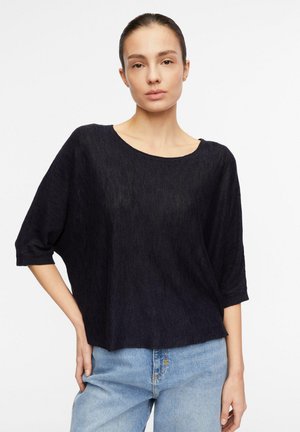 Frau mit dunkel zurückgebundenem Haar, die ein lockeres schwarzes Oberteil mit Dreiviertelärmeln und hellblaue Jeans trägt, steht vor einem weißen Hintergrund.