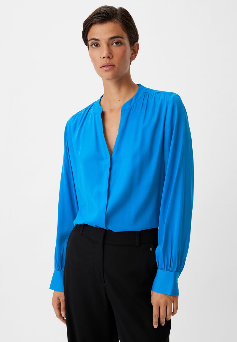Blaue Seidenbluse mit tiefem V-Ausschnitt, langen Ärmeln und gerafften Details an den Schultern, kombiniert mit schwarzen Hosen.