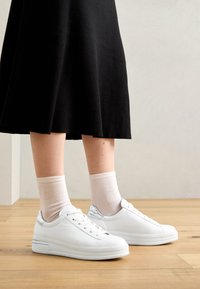 Witte leren sneakers met een gladde textuur, voorzien van een zilveren accent op de hiel, gecombineerd met lichtgekleurde sokken en een zwarte rok.