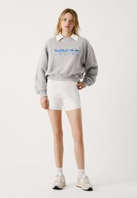 Felpa grigia con logo "Red Bull Racing", colletto bianco, abbinata a pantaloni corti bianchi con accenti laterali blu e sneakers beige.
