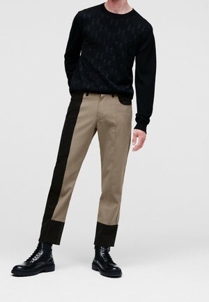 Man met een zwart gebreide trui met patroon, beige broek met zwarte zij- en onderpanelen, en zwarte veterlaarzen, staand tegen een witte achtergrond.