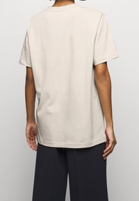 T-shirt beige in cotone a maniche corte, con scollo rotondo e vestibilità comoda, abbinata a pantaloni scuri, vista da dietro.