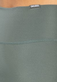 Zelené sportovní šortky vyrobené z elastického materiálu, s hladkým povrchem, plochými švy a černou visačkou "SMILODOX" na pase.