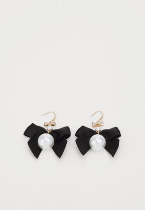 Boucles d'oreilles à crochets en or avec de grosses perles blanches, petits nœuds en cristal et nœuds en tissu noirs sur fond blanc.
