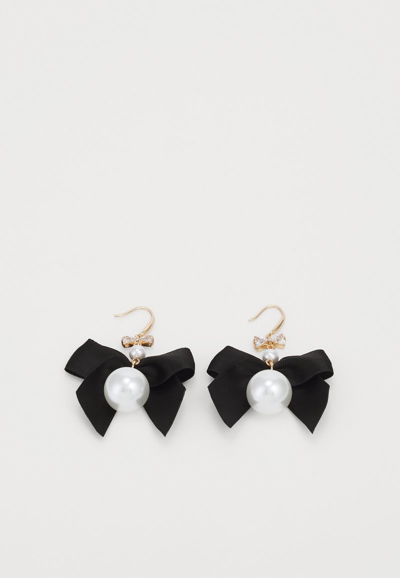 Boucles d'oreilles à crochets en or avec de grosses perles blanches, petits nœuds en cristal et nœuds en tissu noirs sur fond blanc.