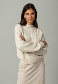 Cremfarbener Kabelstrickpullover mit hohem Kragen, weiten Ärmeln und geripptem Saum; über einem hellbeigen Rock kombiniert. Strukturiertes, gemütliches Aussehen.