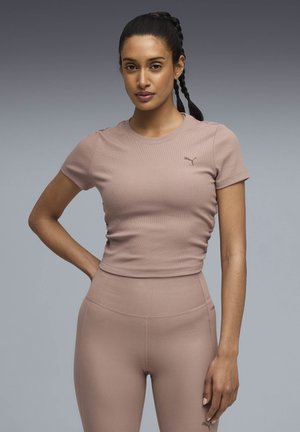 Νεαρή γυναίκα με πλεγμένα μαλλιά, φοράει κορυφή κομμένης γραμμής Puma σε αποχρώσεις του μοβ και κολάν με ψηλή μέση, στέκεται μπροστά σε γκρίζο διαβαθμισμένο φόντο.