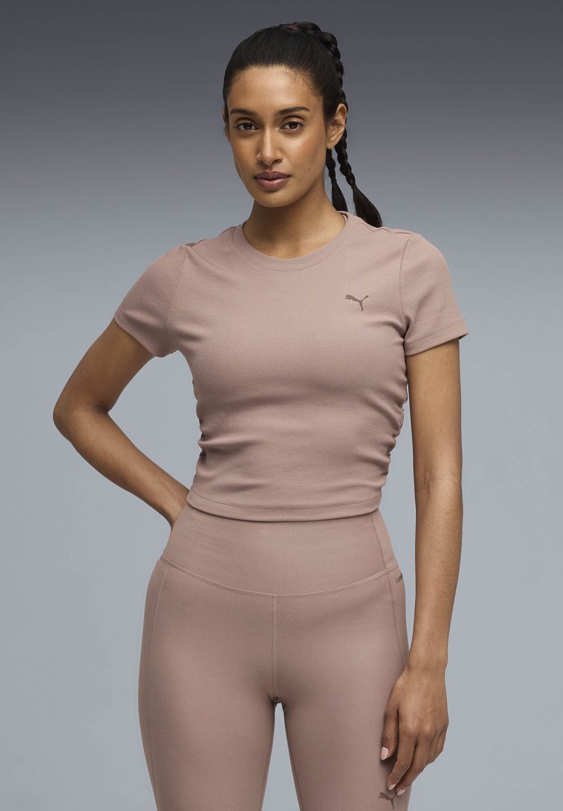 Νεαρή γυναίκα με πλεγμένα μαλλιά, φοράει κορυφή κομμένης γραμμής Puma σε αποχρώσεις του μοβ και κολάν με ψηλή μέση, στέκεται μπροστά σε γκρίζο διαβαθμισμένο φόντο.