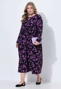 Donna che indossa un vestito midi nero con un motivo floreale viola, tiene in mano una pochette trapuntata lavanda e indossa ballerine nere e orecchini d'argento.