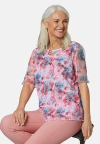 Durchsichtige, kurzärmelige Bluse mit einem pinken Blumenmuster, gewellten Säumen und einem runden Halsauschnitt, kombiniert mit hellpinken Hosen.