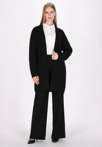 Schwarzer Cardigan mit langen Ärmeln und offener Front, mit einem strukturierten Strickdesign, getragen über einem weißen Kragenhemd und einer schwarzen, weit geschnittenen Hose.
