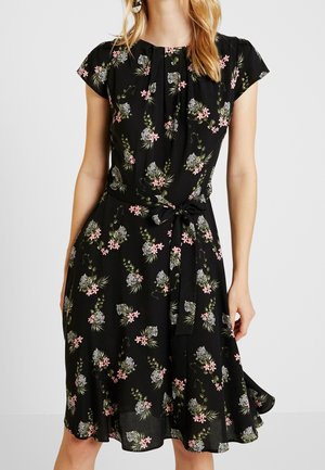 Vrouw draagt een zwarte jurk tot op de knie met korte mouwen, roze bloemmotief en een afbeelding van een witte tijgerkop, en een tailleceintuur die gestrikt is.