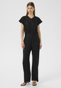 Combinaison en lin noire avec des manches courtes, une taille froncée et des jambes larges. Portée avec des sandales noires et un bracelet en or.