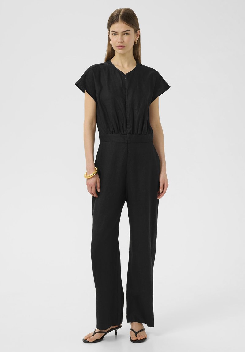 Combinaison en lin noire avec des manches courtes, une taille froncée et des jambes larges. Portée avec des sandales noires et un bracelet en or.