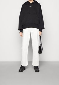 Haut à capuche Nike noir avec un logo blanc, associé à un pantalon blanc ample et des chaussures noires. Un petit sac à main noir complète le look.