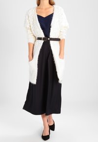 Witte donzige cardigan met oversized zakken, gedragen over een marineblauwe top en zwarte wijde broek, om de taille cinched met een zwarte riem.