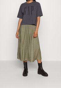 Grijze cropped t-shirt met groot "GAP 1969" logo, gecombineerd met een geplooide olijfgroene rok van gemiddelde lengte en zwarte enkellaarsjes.