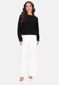 Fekete kötött pulóver hosszú ujjal, fehér bő szárú nadrággal és fehér magassarkúval párosítva, semleges háttér előtt.