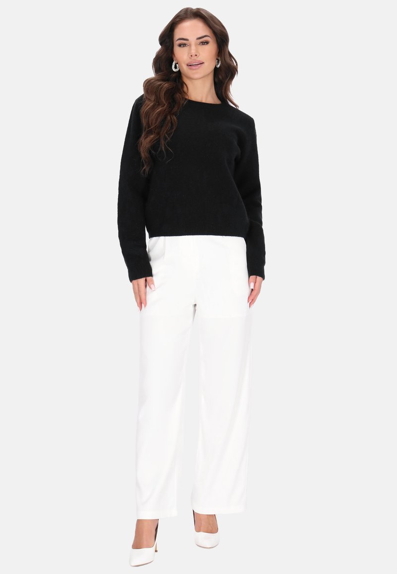Fekete kötött pulóver hosszú ujjal, fehér bő szárú nadrággal és fehér magassarkúval párosítva, semleges háttér előtt.