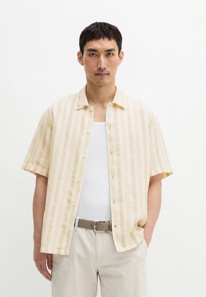 Mann trägt ein beige gestreiftes kurzärmeliges Hemd über einem weißen Tanktop mit beiger Hose und Gürtel, steht vor weißem Hintergrund.