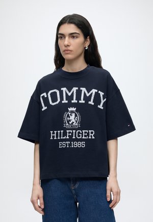 Ung kvinde med langt mørkt hår iført en løs navy Tommy Hilfiger t-shirt med hvidt logo og blå jeans, står foran en ensfarvet baggrund.