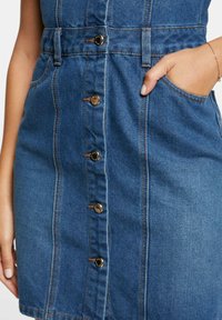 Robe en denim avec un design ajusté, couleur bleu clair, boutons sur le devant et poches latérales. Coutures dans une couleur beige contrastante.